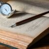 brown pen on top of book beside gray pocket watch